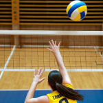 **

A dynamic action shot of a volleyball player mid-air, arms extended high above the net, hands angled inwards forming a blocking "roof." Focus on the intensity in their eyes and the blurred movement of the approaching volleyball. The background shows a brightly lit volleyball court with teammates in supporting positions. Emphasize the power and precision of the block.

**